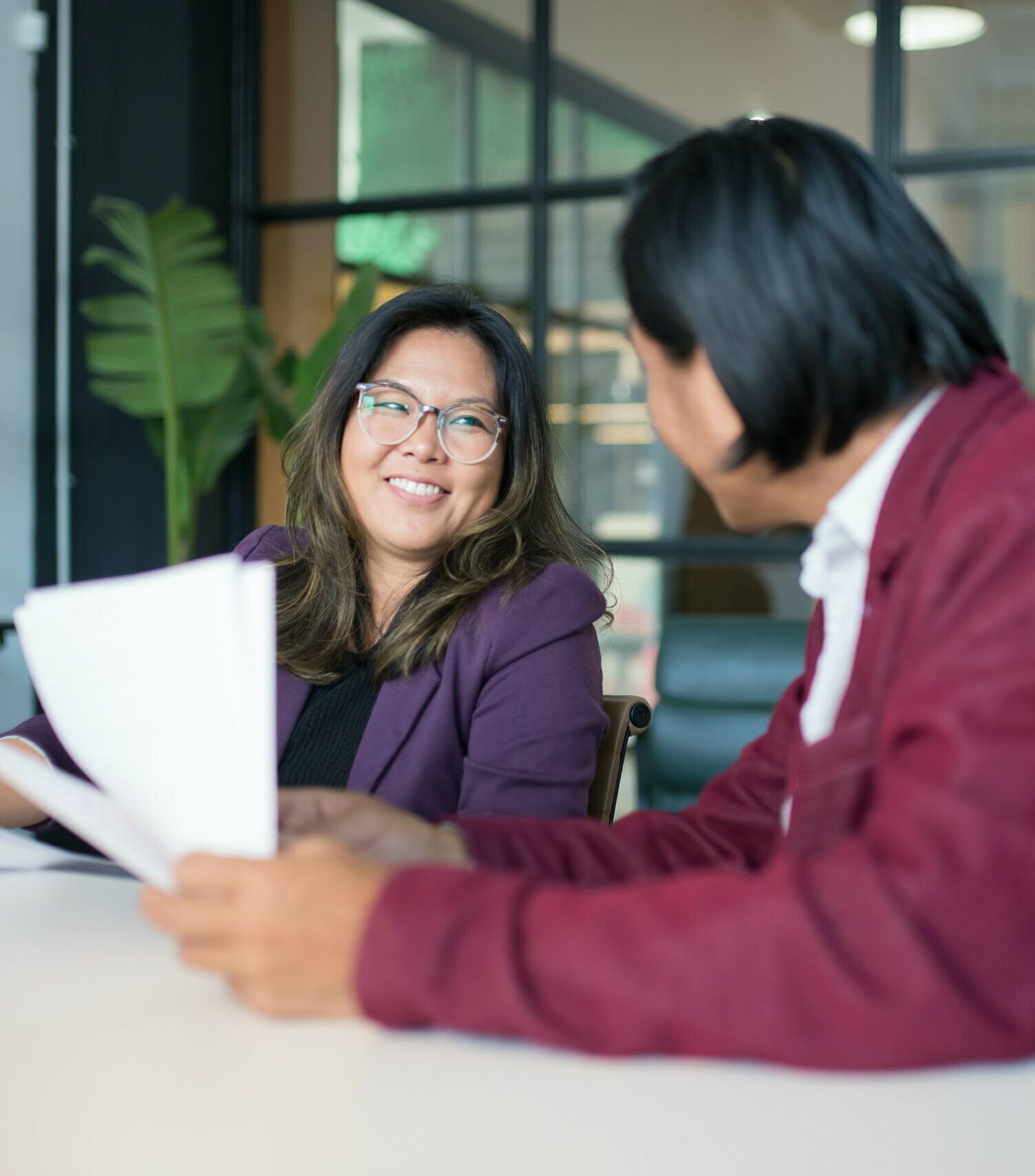 Employees discuss documents in a modern boardroom, smiling sitting at a large table holding papers