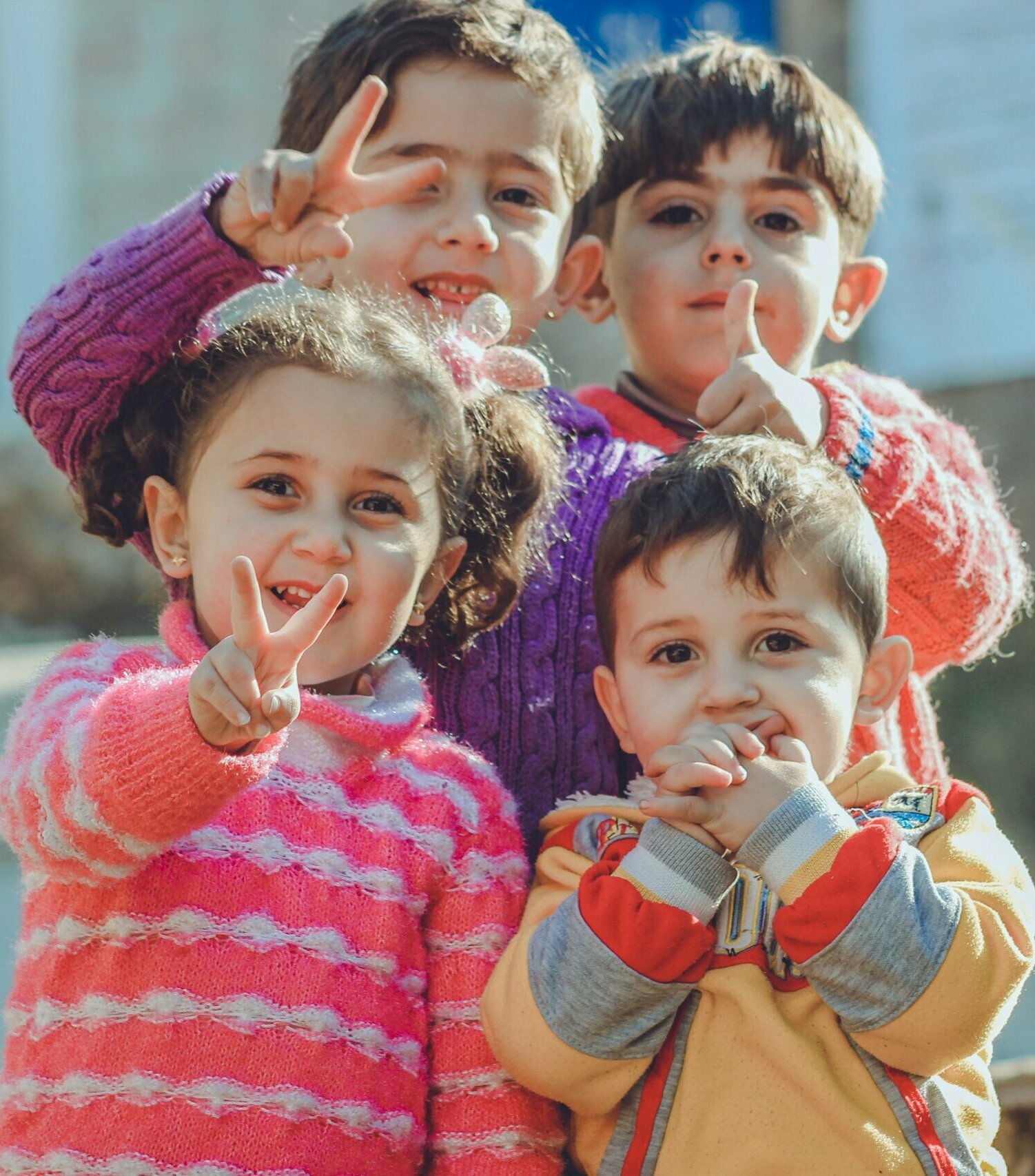 Four young children wearing brightly coloured knitted clothing giving peace signs and thumbs up